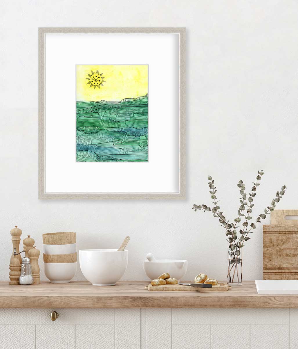 A green and yellow abstract landscape framed in a silver frame with a mat hangs on a white wall above a butcher block countertop with bowls, a cutting board and a clear glass vase with eucalyptus stems.