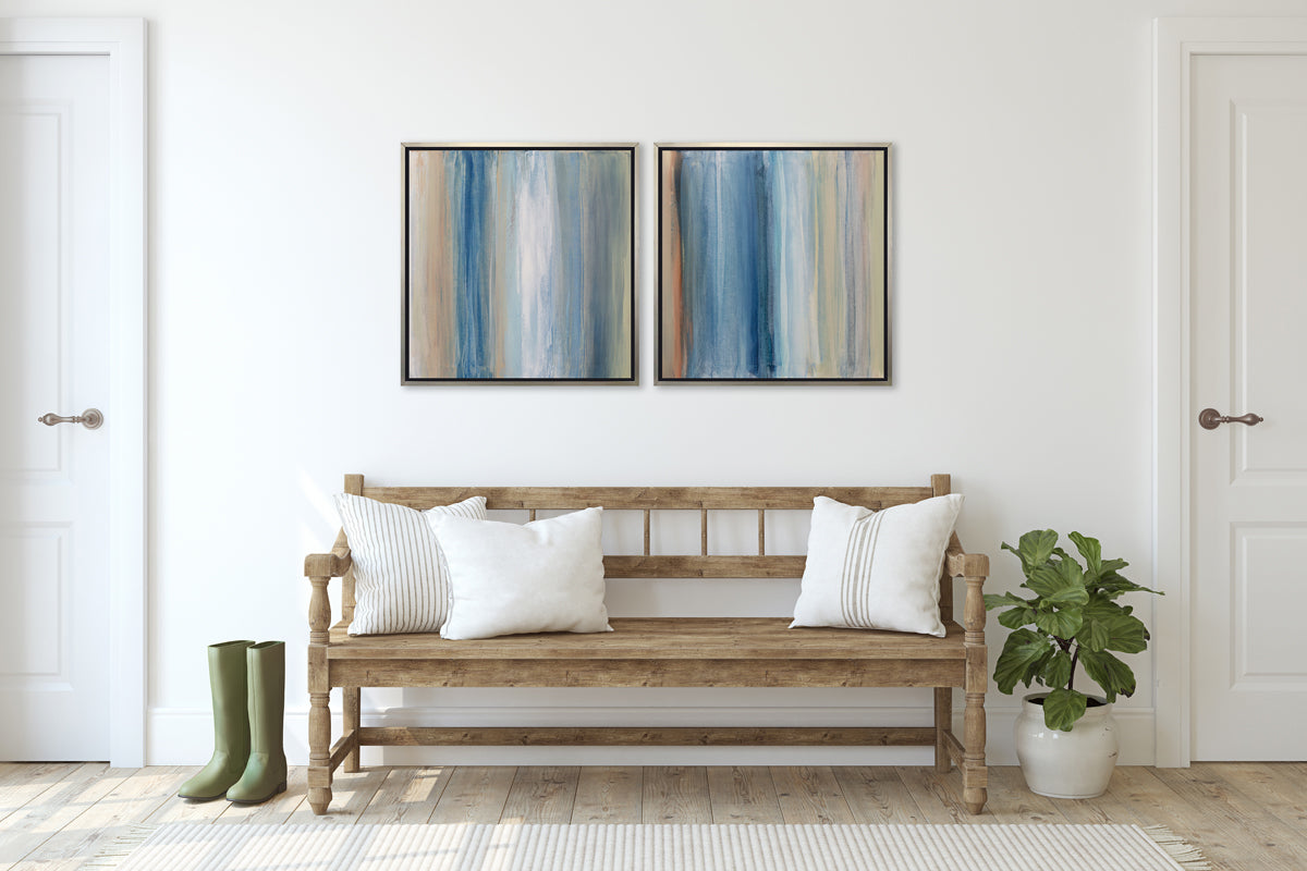 Two blue, white, orange and grey abstract prints in silver floater frames hang on a white wall above a natural wood bench with neutral pillows, a potted floor plant to one side and a pair of green rubber boots to the other.
