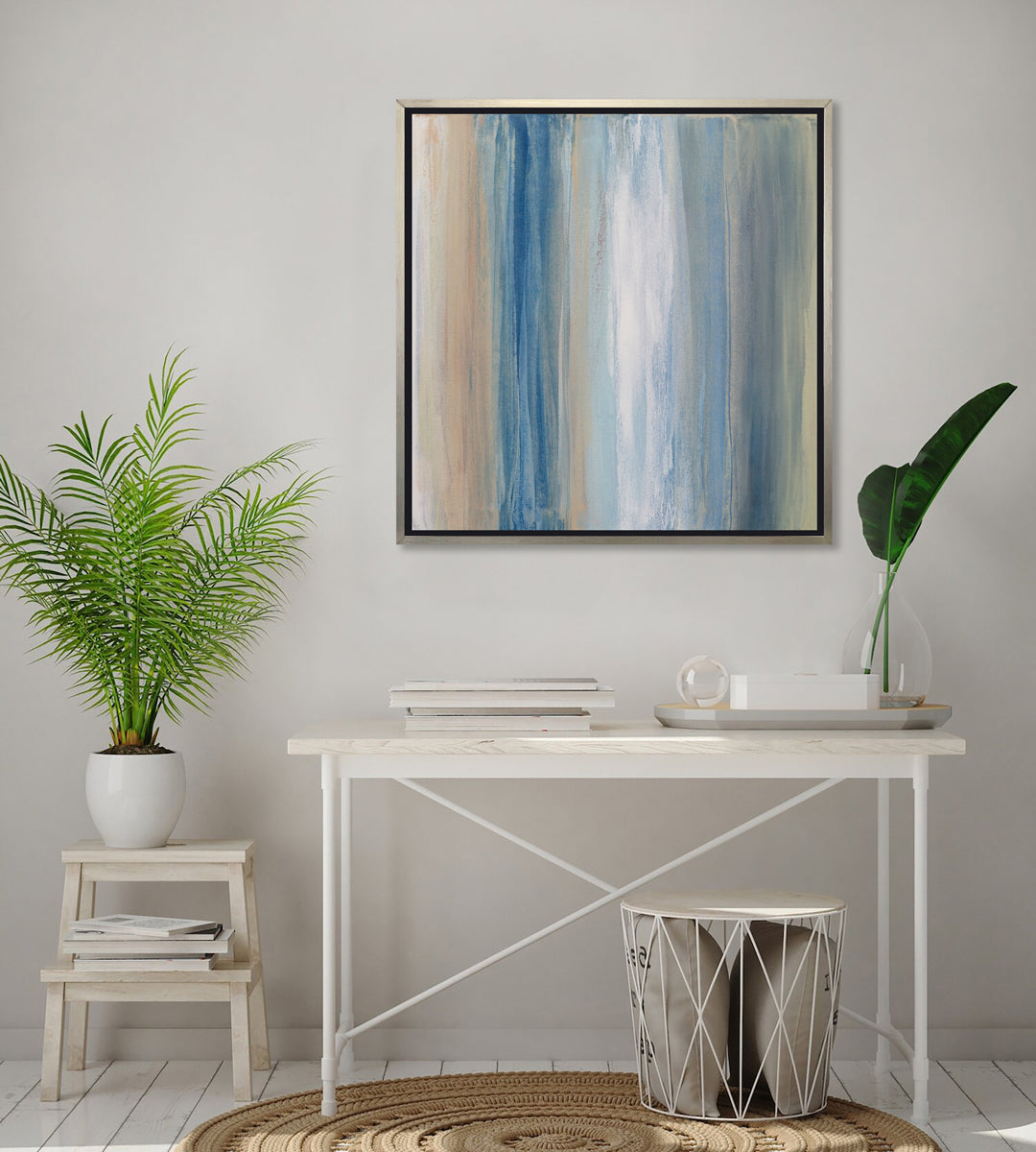 A blue, white, orange and grey abstract print in a silver floater frame hangs on a white wall above a white desk with a stack of books, a tray with decorative items and a step stool with a potted plant.