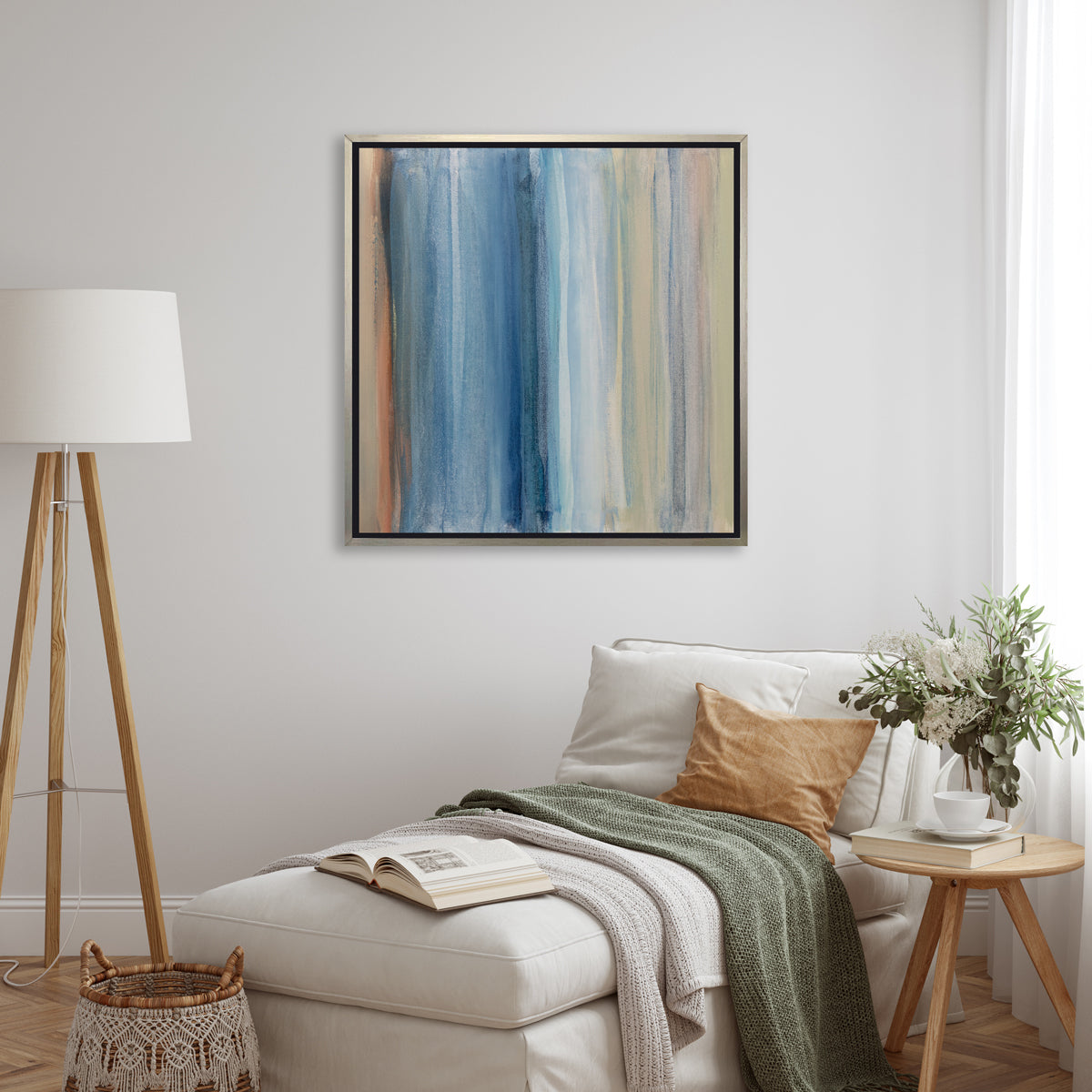 A blue, white, orange and light green abstract print in a silver floater frame hangs on a white wall above a white lounger with pillows and a green throw blanket, a small side table with accessories and a matching tripod floor lamp.