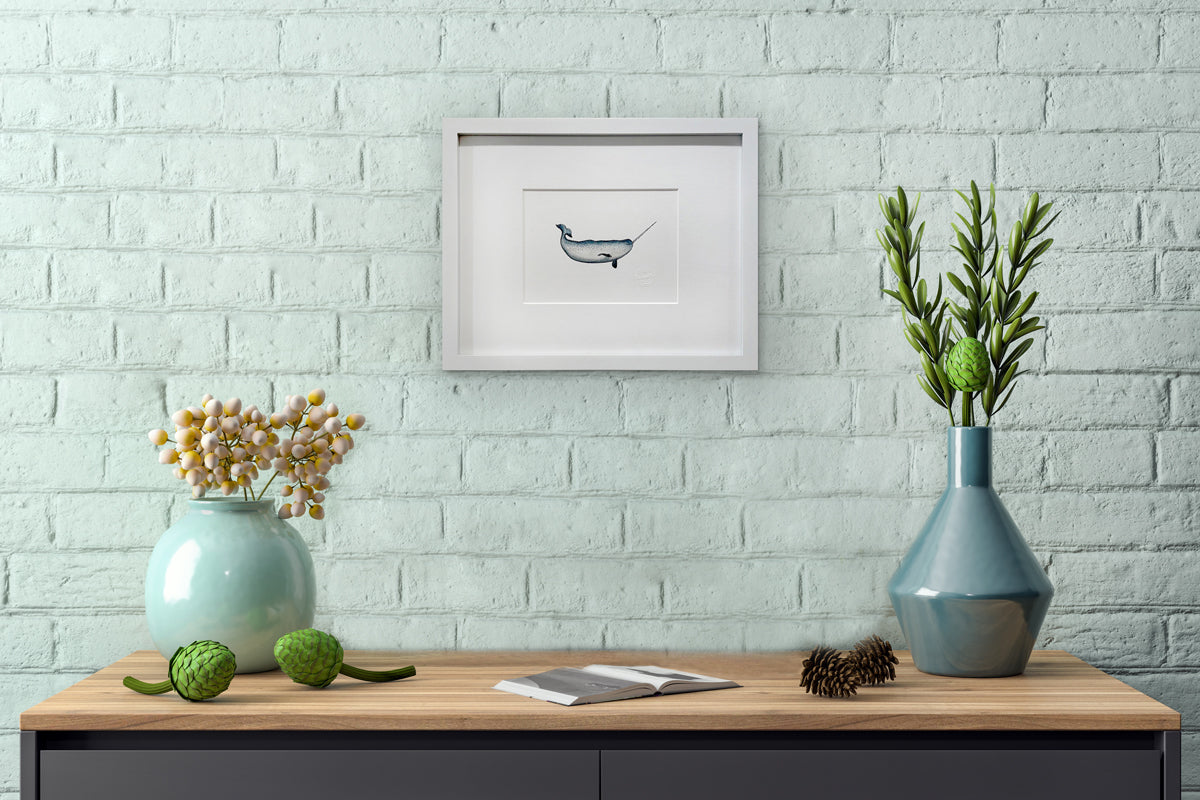 A blue, white and black illustration drawing of a narwhal sea creature in a white frame with a mat hangs on a light teal brick wall above a wood surface with an open book and two teal vases with succulent arrangements.