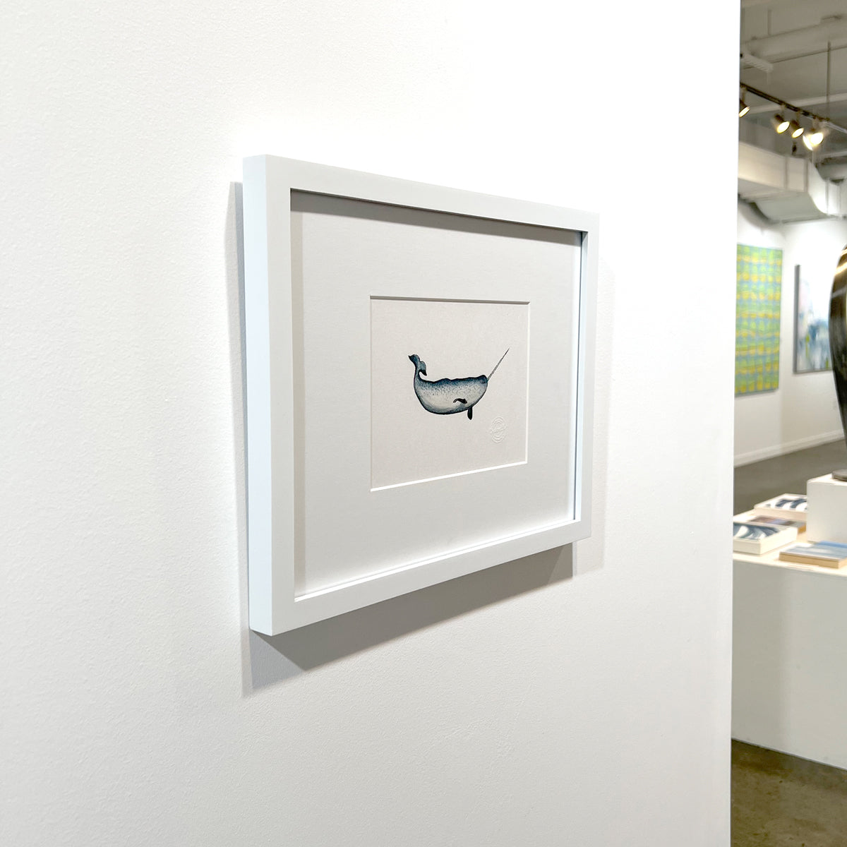 A left side-angle view of a blue, white and black illustration drawing of a narwhal sea creature in a white frame with a mat hangs on a white wall.