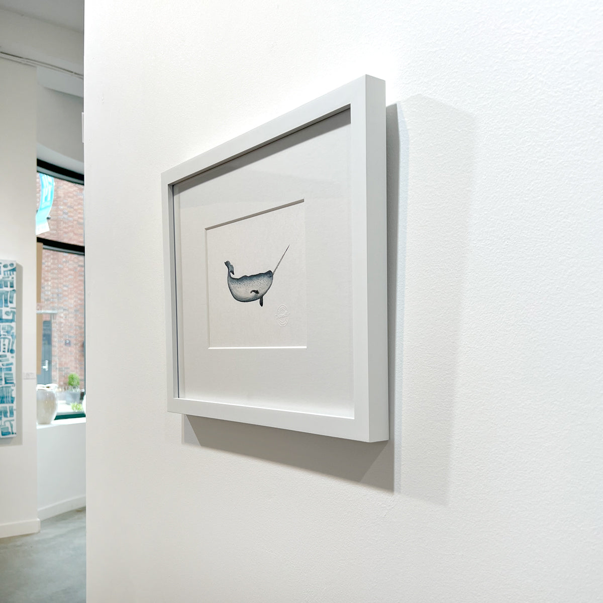 A right side-angle view of a blue, white and black illustration drawing of a narwhal sea creature in a white frame with a mat hangs on a white wall.