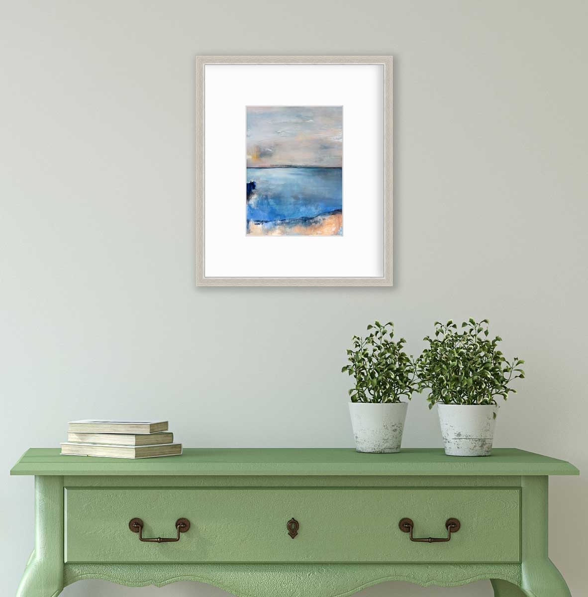 A blue and grey abstract print in a silver frame with a mat hangs on a white wall above a green console table with a stack of books and two potted plants.