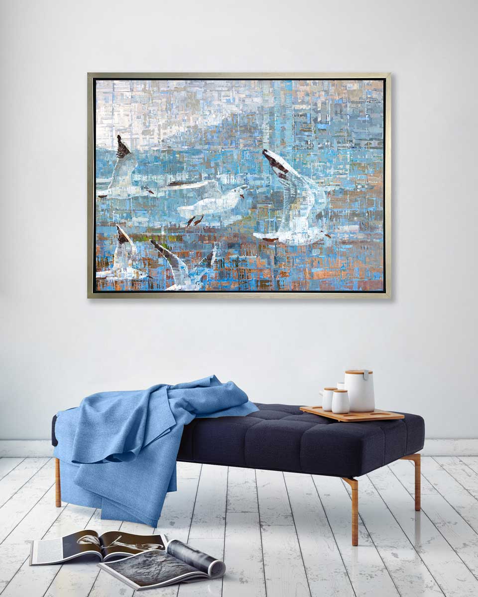 A teal and white geometric abstract print of birds flying together in a silver floater frame hangs on a white wall above a dark blue ottoman with a light blue throw blanket, a natural wood tray with white containers and two open magazines on the floor.