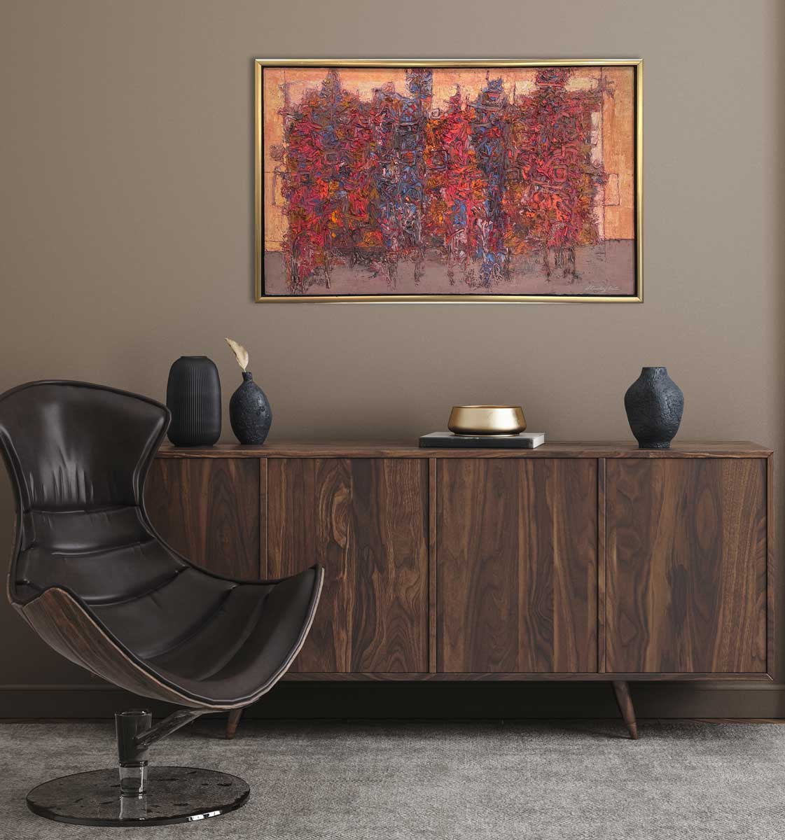 A multicolor abstract painting in a warm gold frame hangs on a grey wall above a stained wood credenza with three black vases, a book with a gold decorative bowl and a brown modern chair.