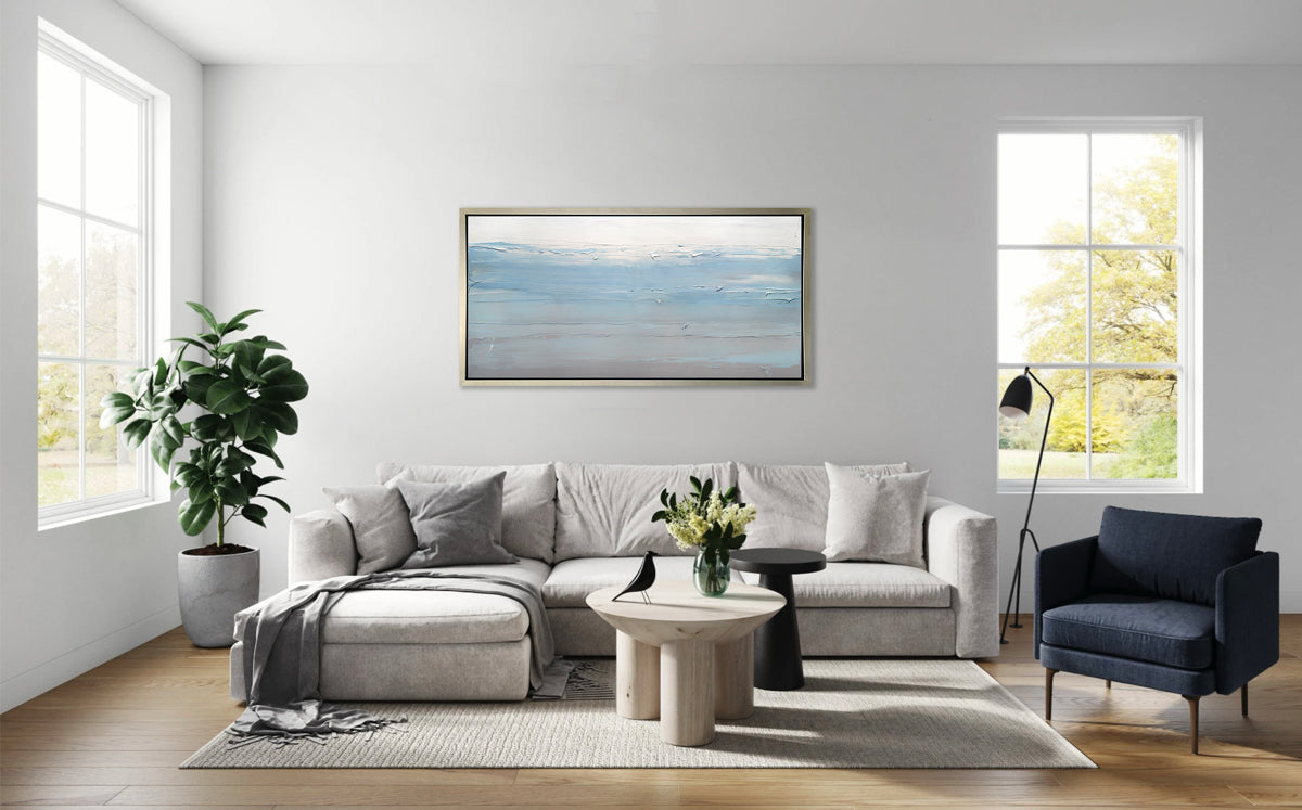 A blue, grey and grey-beige abstract print in a silver floater frame hangs on a white wall above a light grey sectional sofa with pillows, a dark navy chair, two rounded coffee tables and a tall potted floor plant.