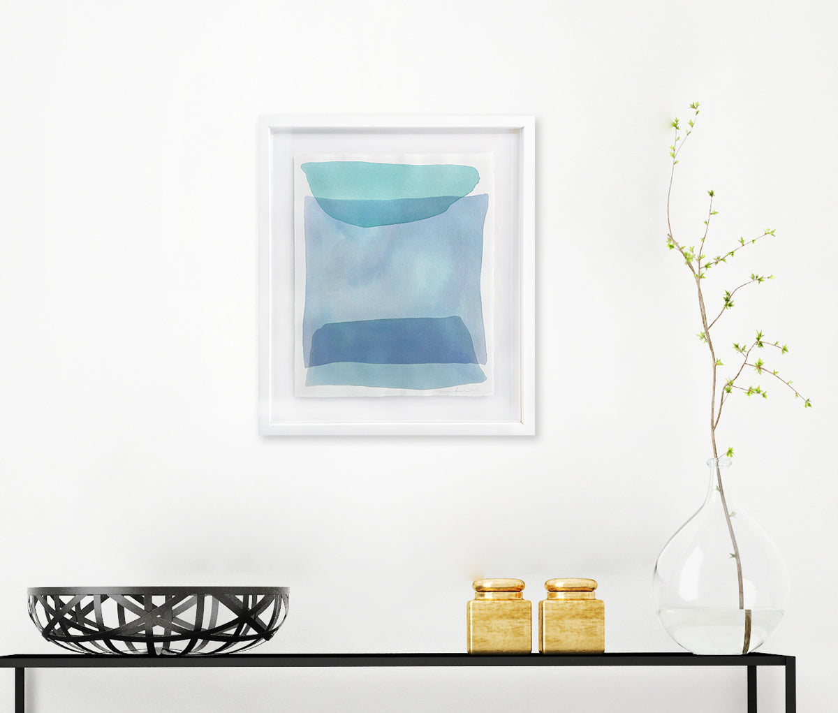 Blue and green shapes overlap in this painting by Nealy Hauschildt in a white frame floating above a white matte hangs on a white wall above a black bookshelf with a black wire bowl, gold accessories and a glass vase with a greenery stem.