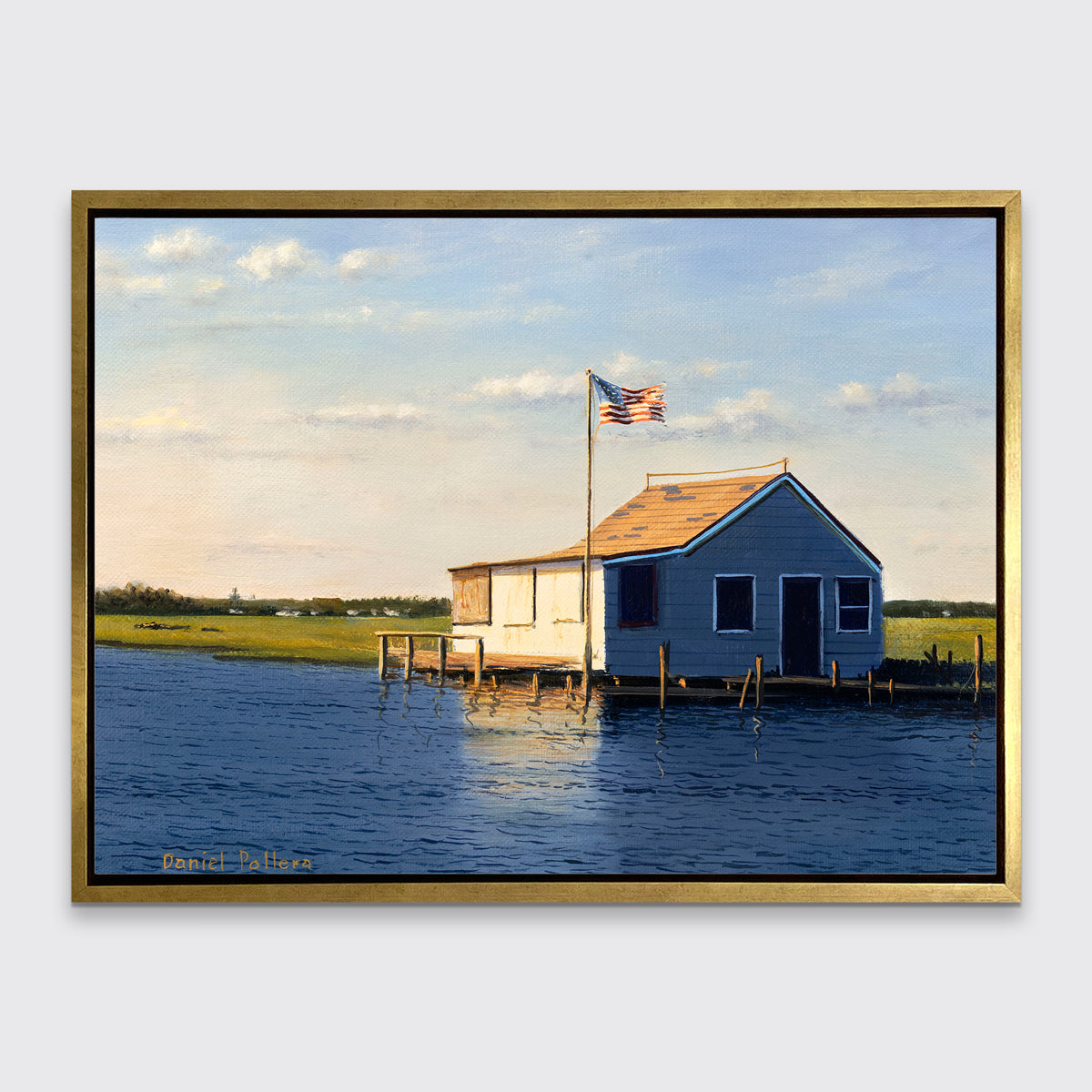 A blue, beige and green contemporary seascape print with a bay house in a gold floater frame hangs on a white wall.