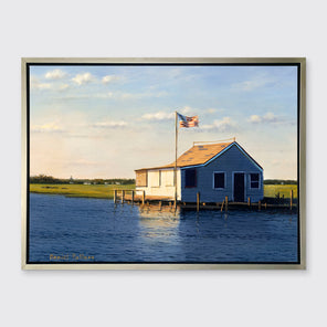 A blue, beige and green contemporary seascape print with a bay house in a silver floater frame hangs on a white wall.