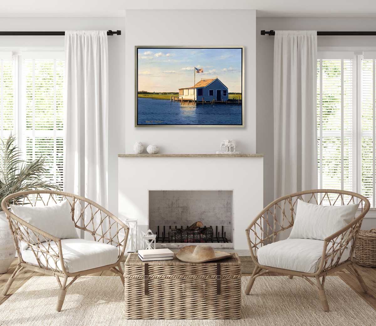 A blue, beige and green contemporary seascape print with a bay house in a silver floater frame hangs on a white wall above a fireplace, two woven chairs with white cushions and a wicker trunk.