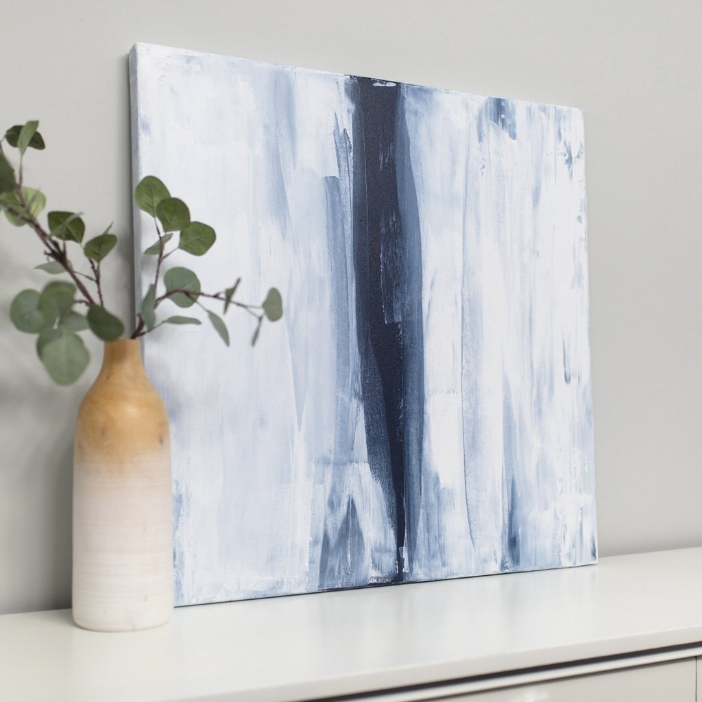 White and blue abstract painting by Julia Contacessi sits on a white dresser leaning against a light grey wall next to a potted plant, viewed from the left. 