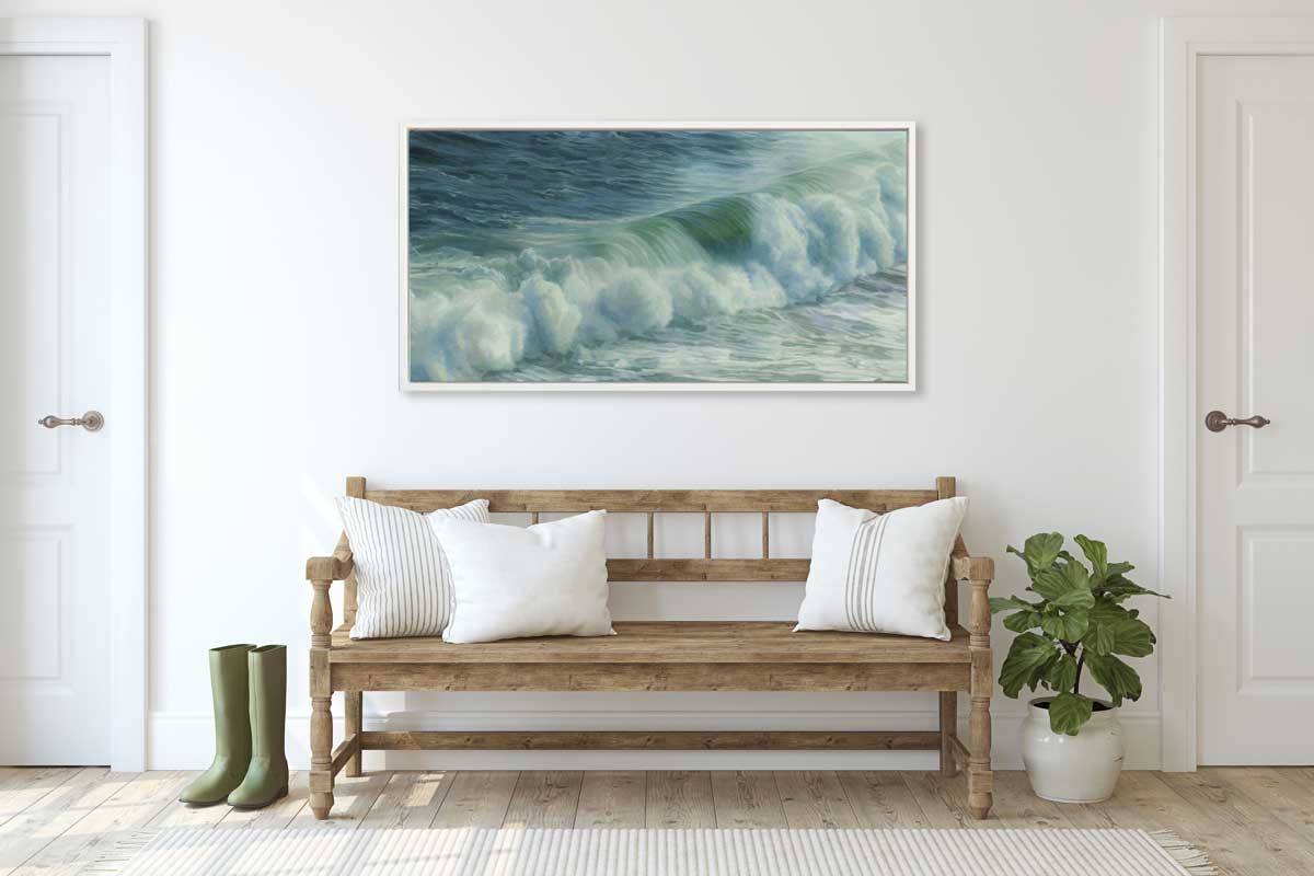 A blue and green abstract water wave print in a white floater frame hangs on a white wall above a wooden bench with white pillows, a pair of green rubber boots and a small potted floor plant.