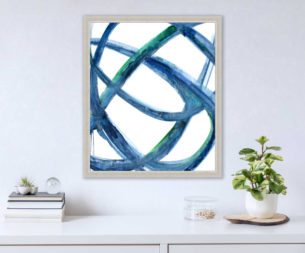 A blue, green and white abstract print in an unmatted silver frame hangs on a white wall above a white surface with a stack of books, a clear round container with gold accessories and a small potted plant.