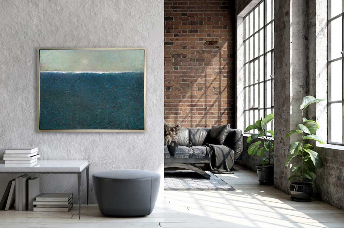 A blue abstract landscape print in a silver floater frame hangs on a white textured side wall above a low white table with books and a grey ottoman. To the right is the opening to a brown leather couch, a glass coffee table and large full wall windows.