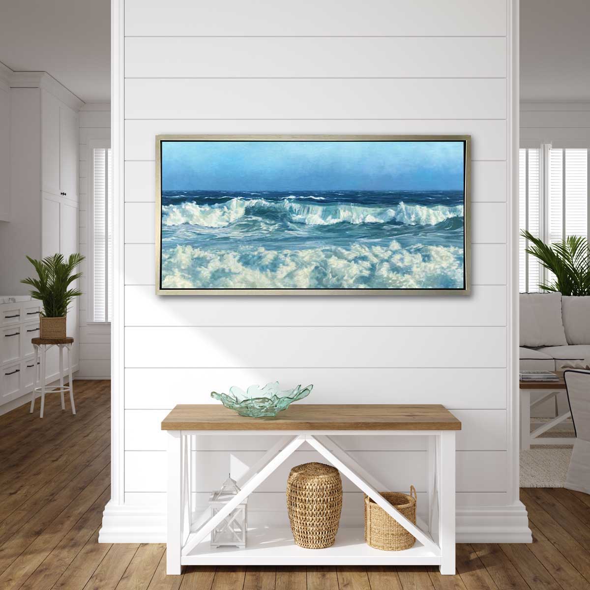 A Blue seascape print in a silver floater frame hangs on a white shiplap wall above a low console table with a green translucent art bowl, a white lantern and two woven baskets.