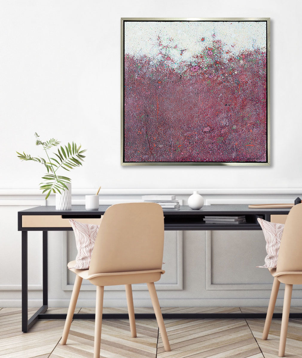 A dark red abstract landscape print in a silver floater frame hangs on a white wall above a large black desk with accessories and two tan chairs with printed pillows.