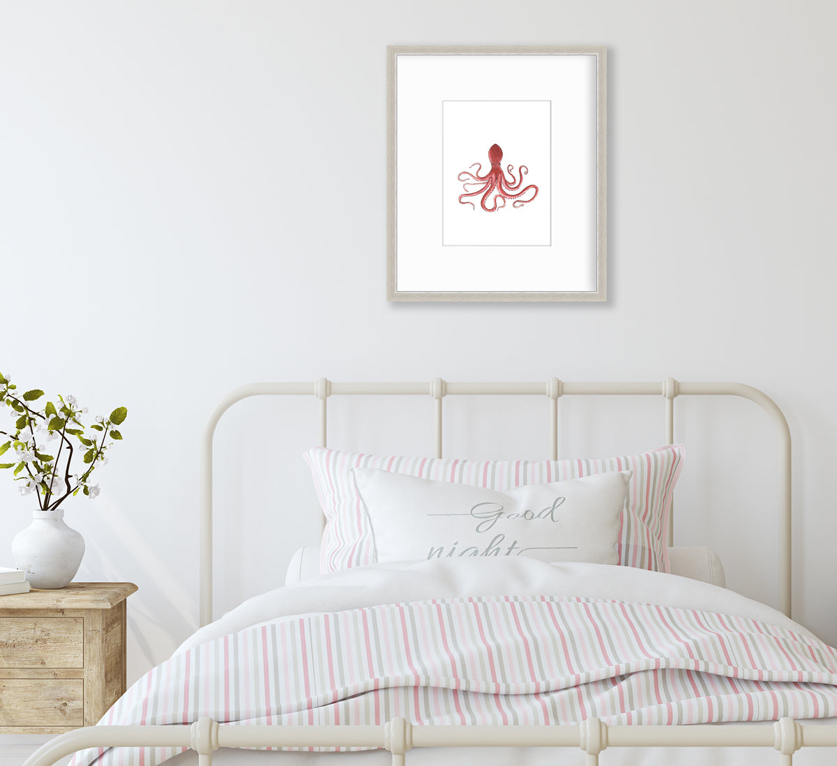 A pink octopus print in a silver frame with a mat hangs on a white wall above a white iron bed with pink and white bedding and a side table with a white vase with greenery stems.