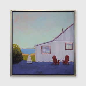A blue, lavender and light purple contemporary seascape print with a beach house, two red adirondack chairs and a walkway leading to the water in a silver floater frame hangs on a white wall.