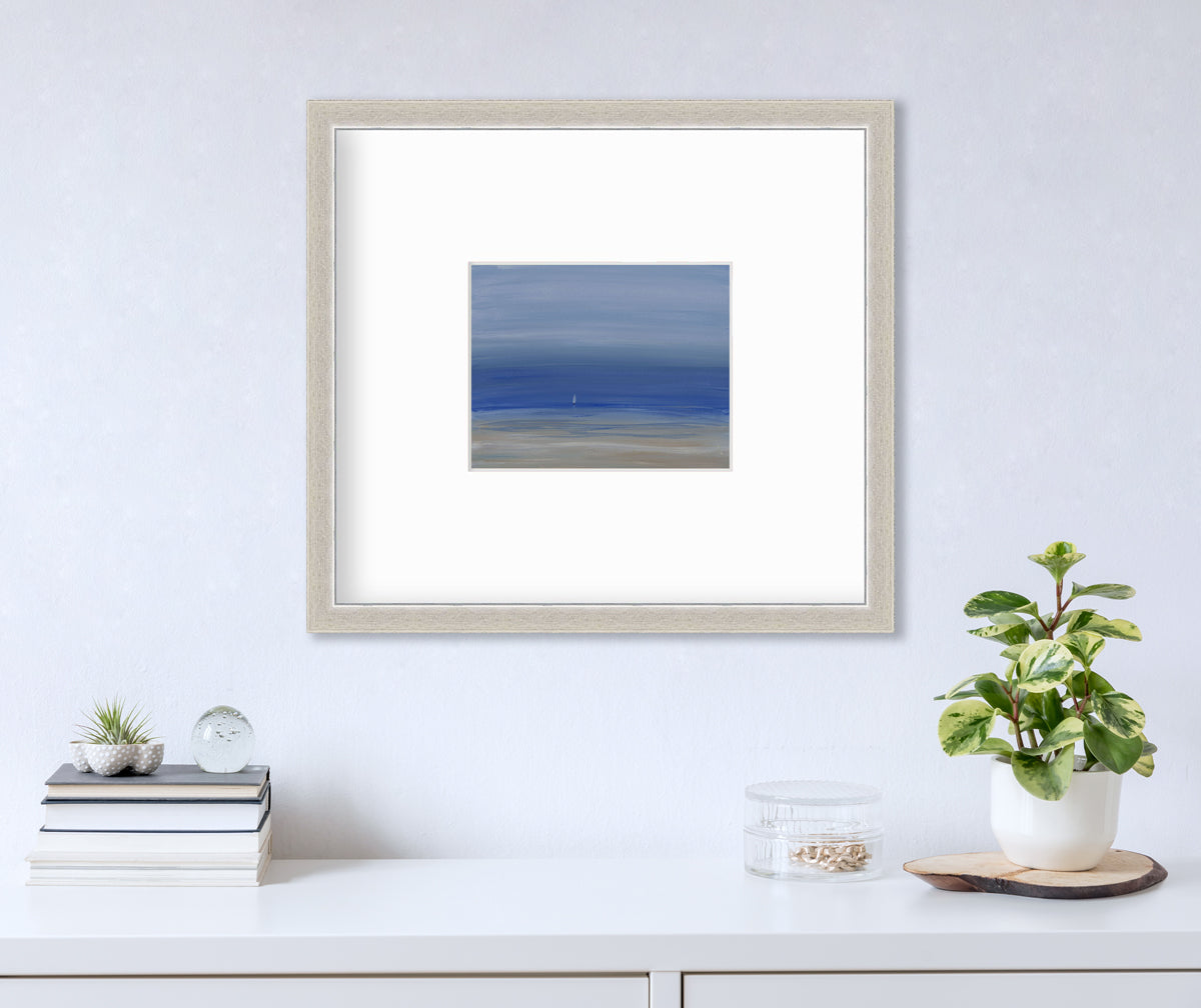 A blue and beige abstract seascape print in a silver frame with a mat hangs on a white wall above a white surface with a stacks of books, a clear round container with gold accessories and a potted plant.