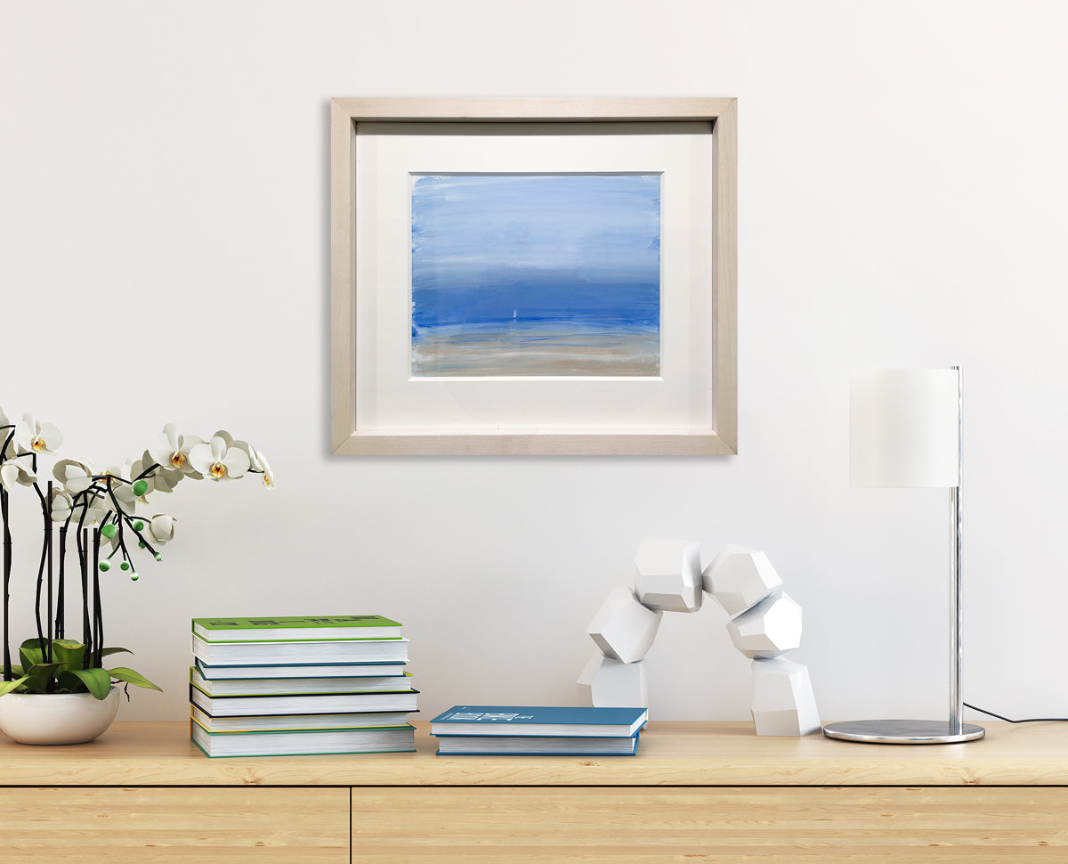A tonal blue and beige abstract coastal seascape with a white sailboat by S. Cora Aldo, is matted and framed in a natural wood frame, hanging on a white wall above a wood surface with stacks of books and decorative items.