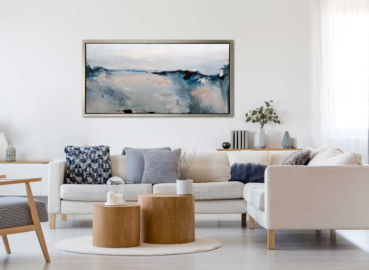A blue, black, grey and peach abstract print in a silver floater frame hangs on a white wall above a white sectional sofa with printed pillows and two round wood coffee tables