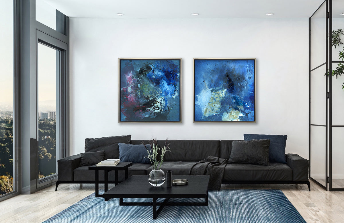 Two blue abstract prints in silver floater frames hang on a white wall above a large black sofa with pillows and a two tier black coffee table.