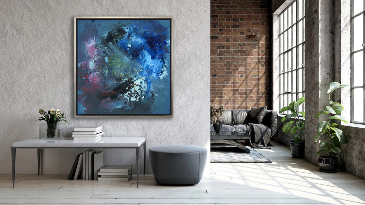 A blue, red, black and green abstract print in a silver floater frame hangs on a grey wall above a white table with a stack of books, a clear vase with a white Lilly arrangement, a dark grey ottoman and next to an opening to a well lit living room.