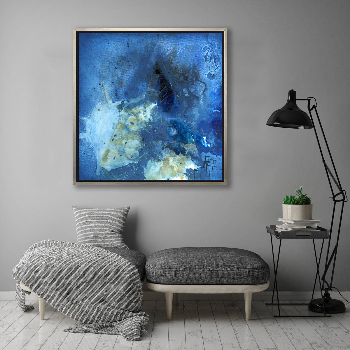A blue, green and black abstract print in a silver floater frame hangs on a grey wall above two grey ottomans, a small side table with a potted succulent arrangement and a black floor lamp.