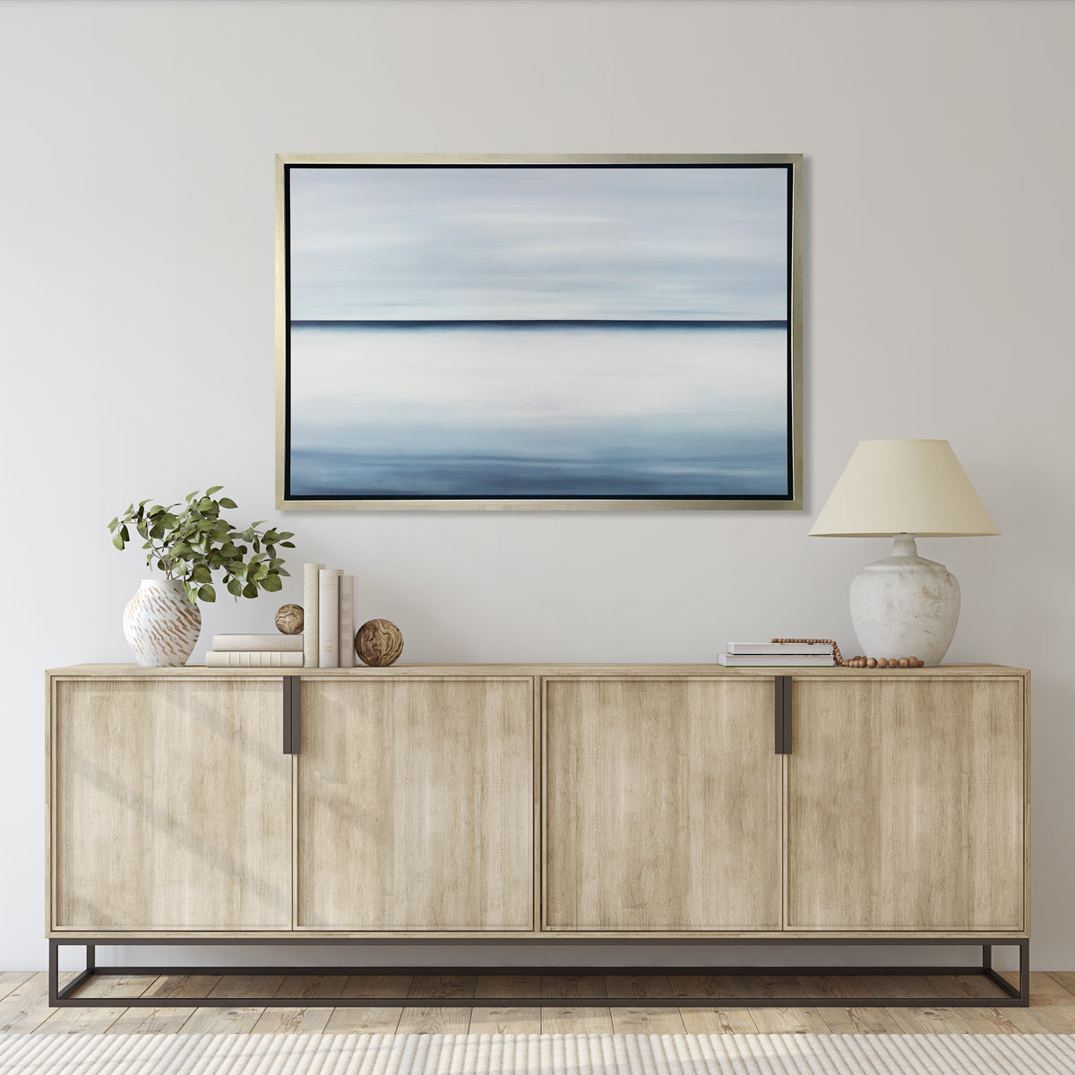 A tonal blue abstract landscape print in a silver floater frame hangs on a white wall above a natural wood credenza with a stack of books, a eucalyptus filled white and beige vase and a large cream colored table lamp.