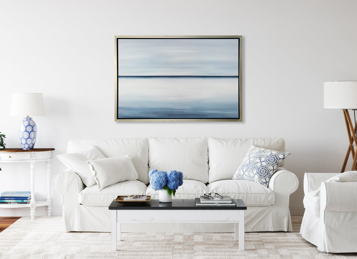 A tonal blue abstract landscape print in a silver floater frame hangs on a white wall above a white sofa with white and blue pillows and a white and black coffee table with decorative accessories