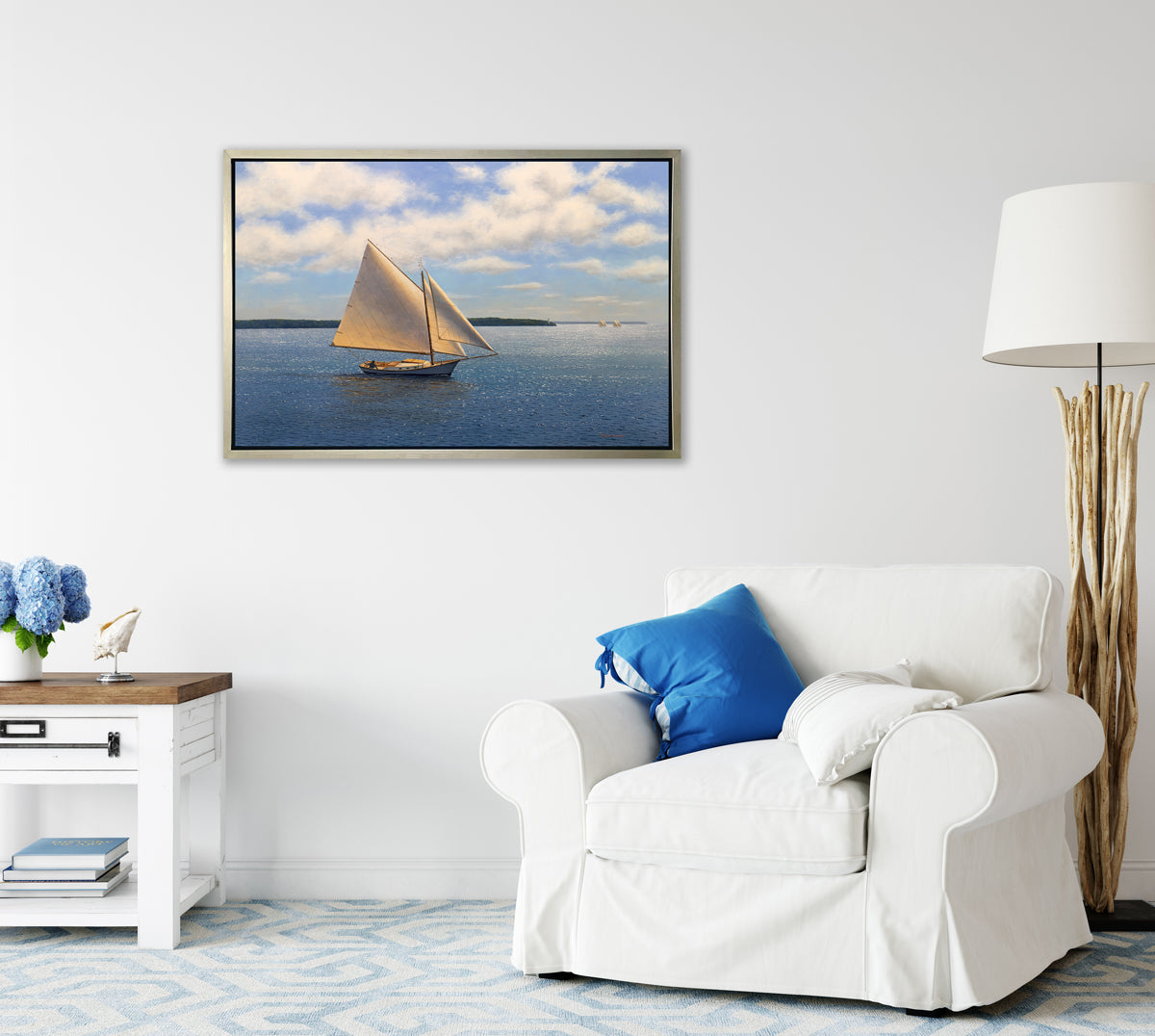 A blue, beige and white seascape print with sailboats in a silver floater frame hangs on a white wall above a white oversized chair with pillows, a white side table and a driftwood floor lamp.