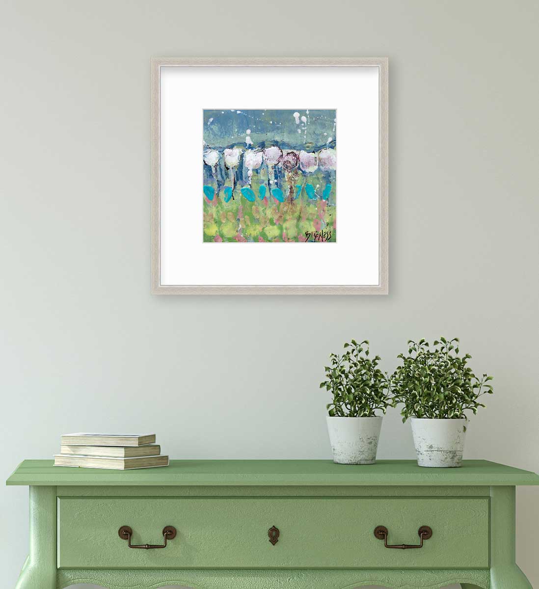 A teal, green and white abstract floral print in a silver frame with a mat hangs on a white wall above a green console table with a stack of books and two potted plants.