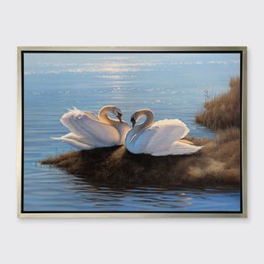 A print of two swans facing each other on an embankment next to water in a silver floater frame hangs on a white wall.