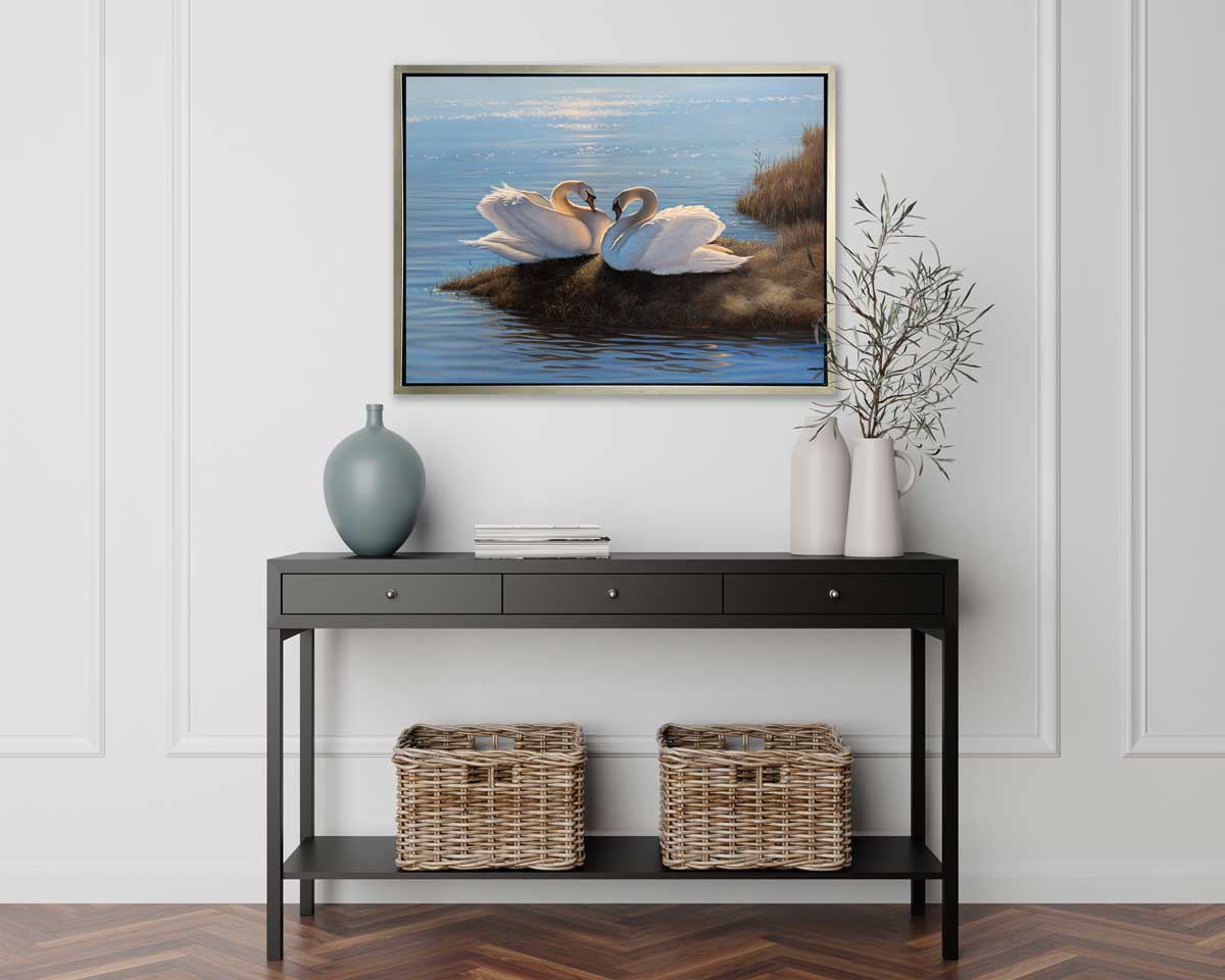 A print of two swans facing each other on an embankment next to water in a silver floater frame hangs on a white wall above a black console table with two white vases with greenery stems, a muted teal vase and two woven baskets.