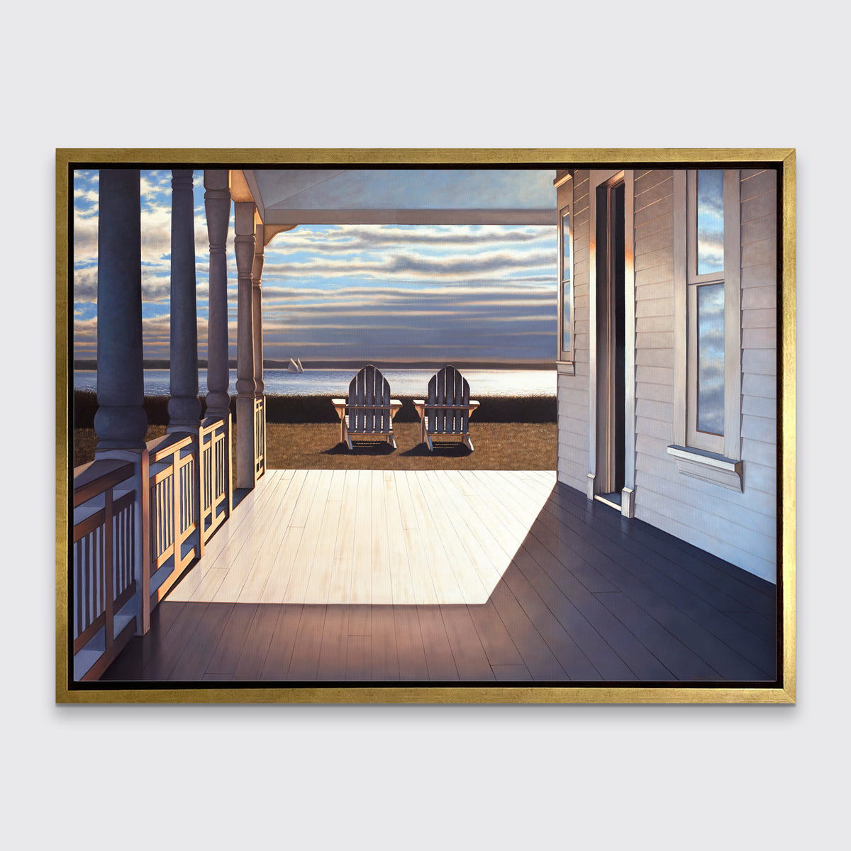 A print of the sunlit porch of a house and two adirondack chairs overlooking the water in a gold floater frame hangs on a white wall.