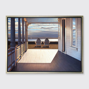 A print of the sunlit porch of a house and two adirondack chairs overlooking the water in a silver floater frame hangs on a white wall.