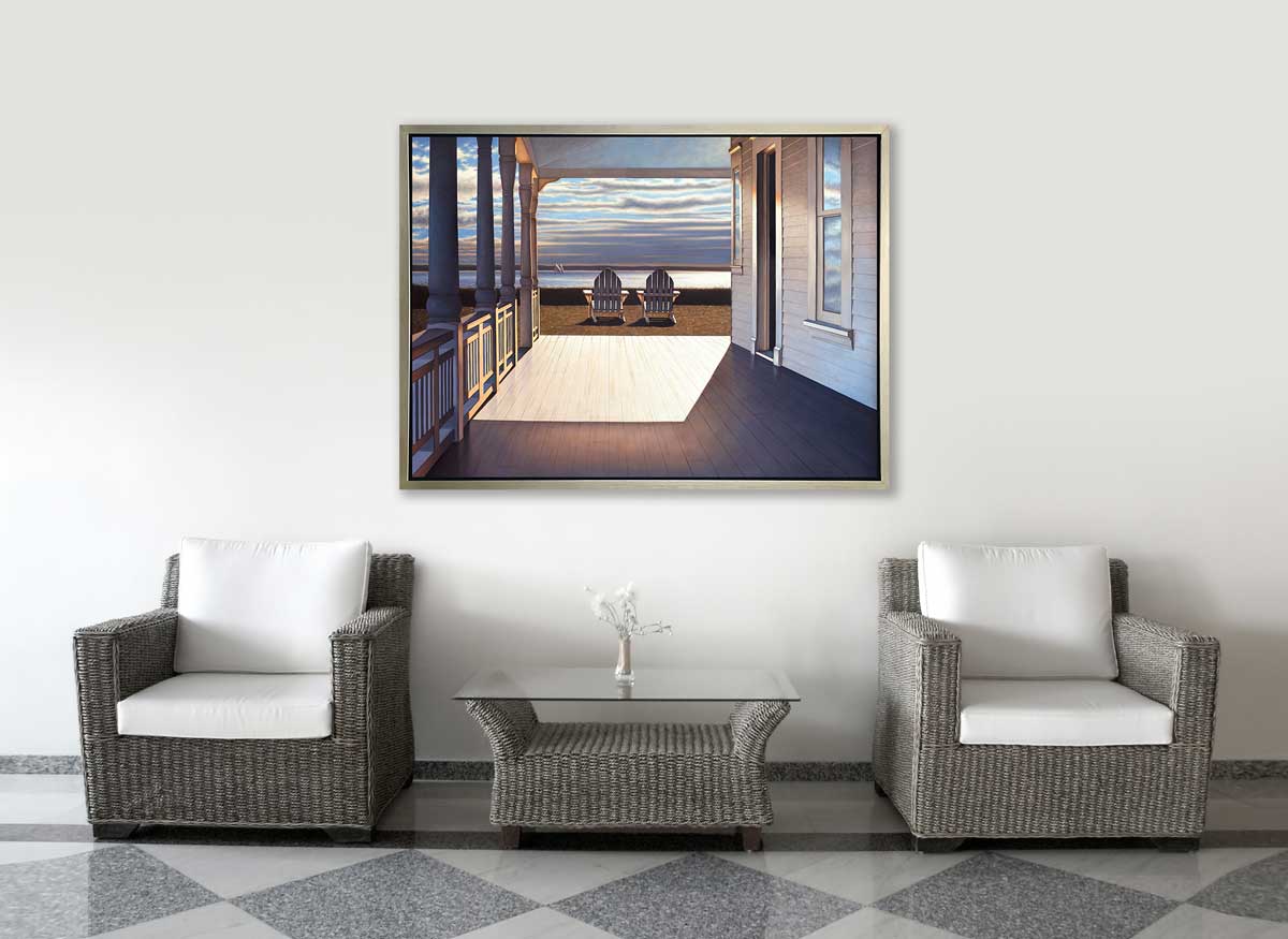 A print of the sunlit porch of a house and two adirondack chairs overlooking the water in a silver floater frame hangs on a white wall above two wicker chairs with white cushions and a wicker coffee table with a glass top.