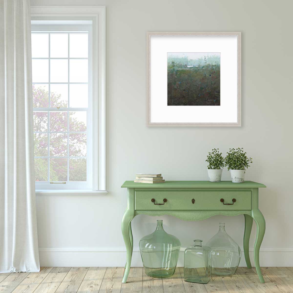 A dark muted green abstract landscape print in a silver frame with a mat hangs on a white wall above a green console table with a stack of books, two potted plants and three green glass bottles.