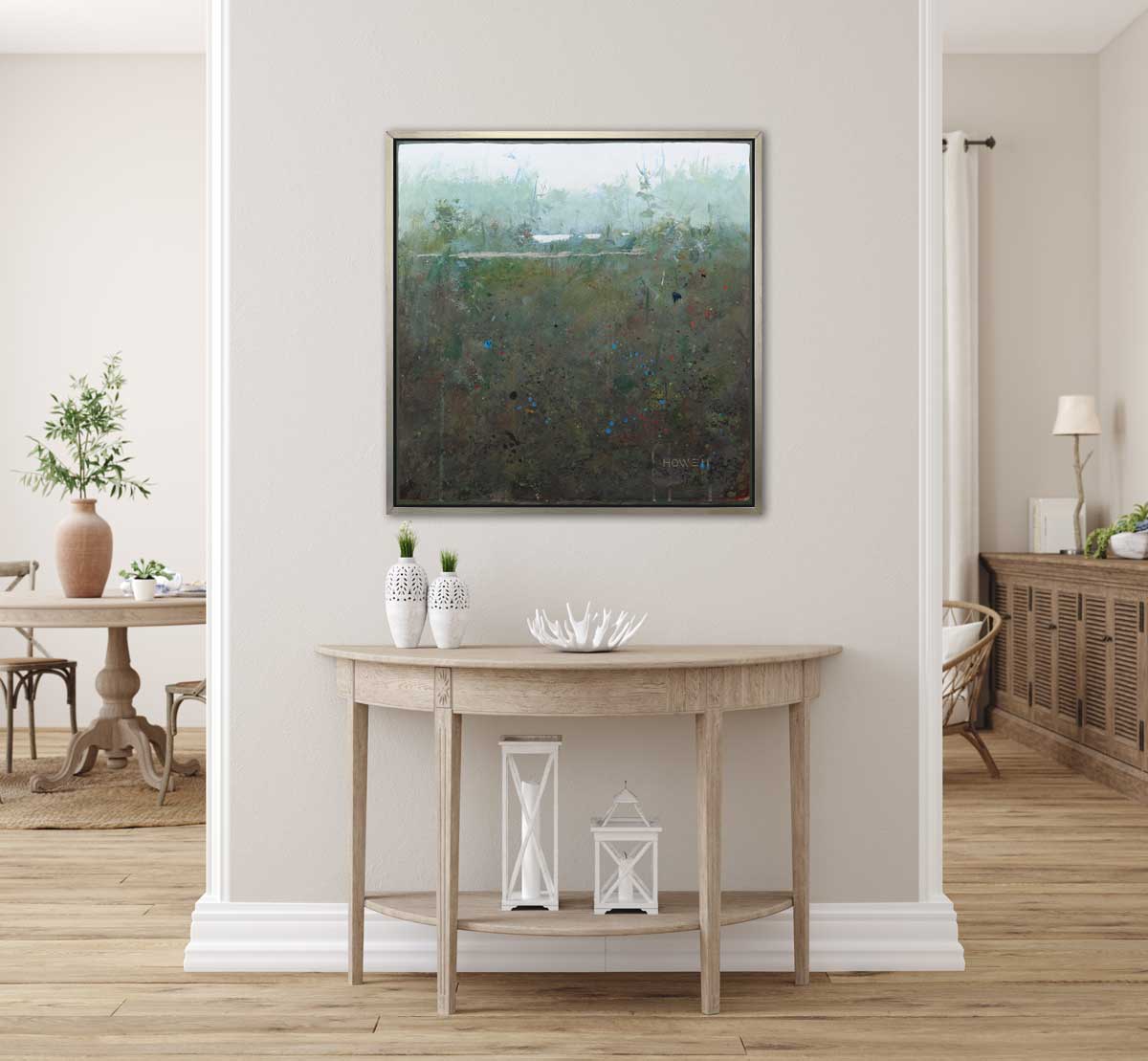 A dark green and grey abstract landscape print in a silver floater frame hangs on a white wall above a natural wood half circle console table with two white vases, a white decorative bowl and two white lanterns.