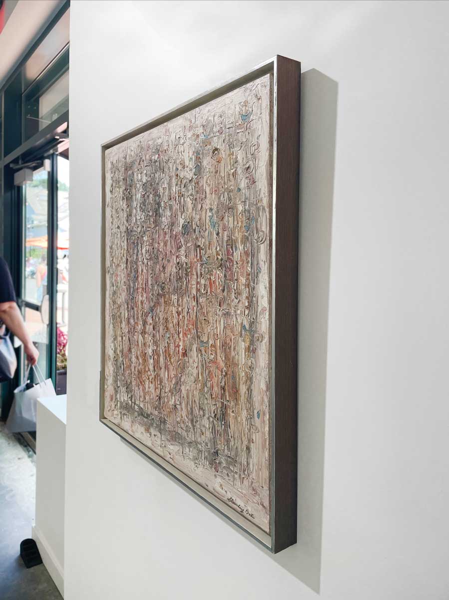 A right side-angle view of a white and multicolored abstract painting in a silver floater frame hangs on a white wall.