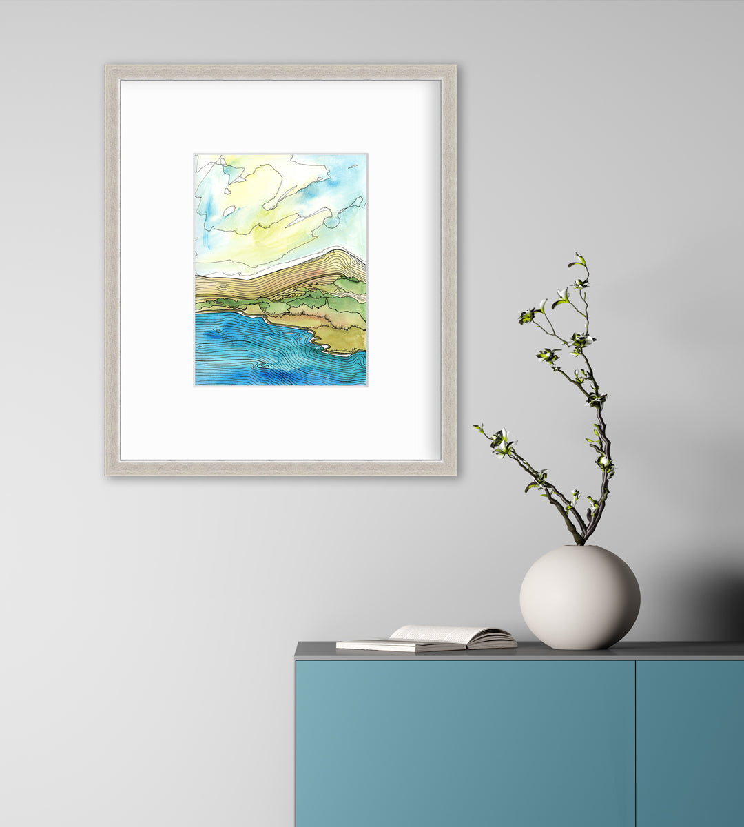A blue, green, brown and light yellow abstract landscape with black outlines print in a silver frame with a mat hangs on a white wall above a teal cabinet with an open book and a white round vase with greenery stems.