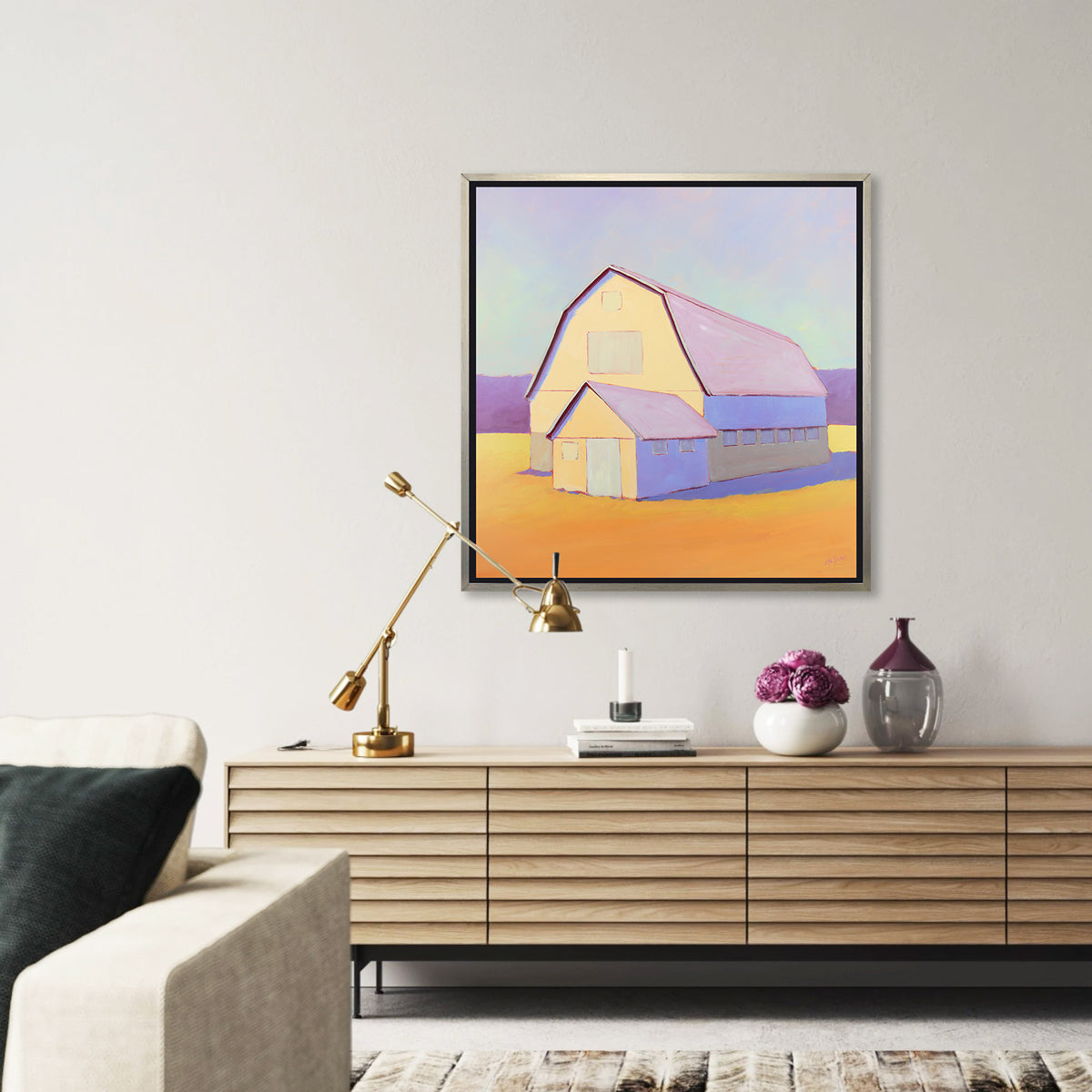 A lavender, light yellow and orange landscape print of a barn in a silver floater frame hangs on a white wall above a natural wood credenza with an adjustable gold lamp, a stack of books and a white vase with pink flowers. 