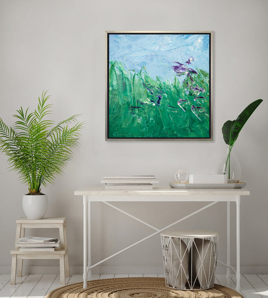 A blue, green and purple abstract landscape print in a silver floater frame hangs on a white wall above a whitewashed desk with a stack of books and a tray with a glass vase with giant leaf stems, a small white step stool that holds a potted fern and a wire storage stool with grey pillows.