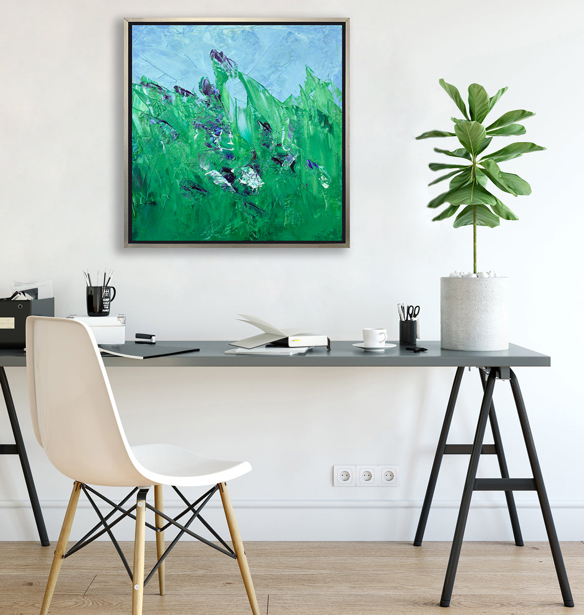 A blue and green abstract landscape print in a silver floater frame hangs on a white wall above a grey desk with open notebooks, black mugs with pencils and a potted plant.