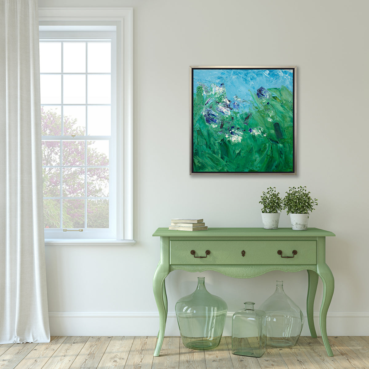 A blue, green, purple and white abstract landscape print in a silver floater frame hangs on a white wall above a green console table with a stack of books, two potted plants and three green glass bottles.