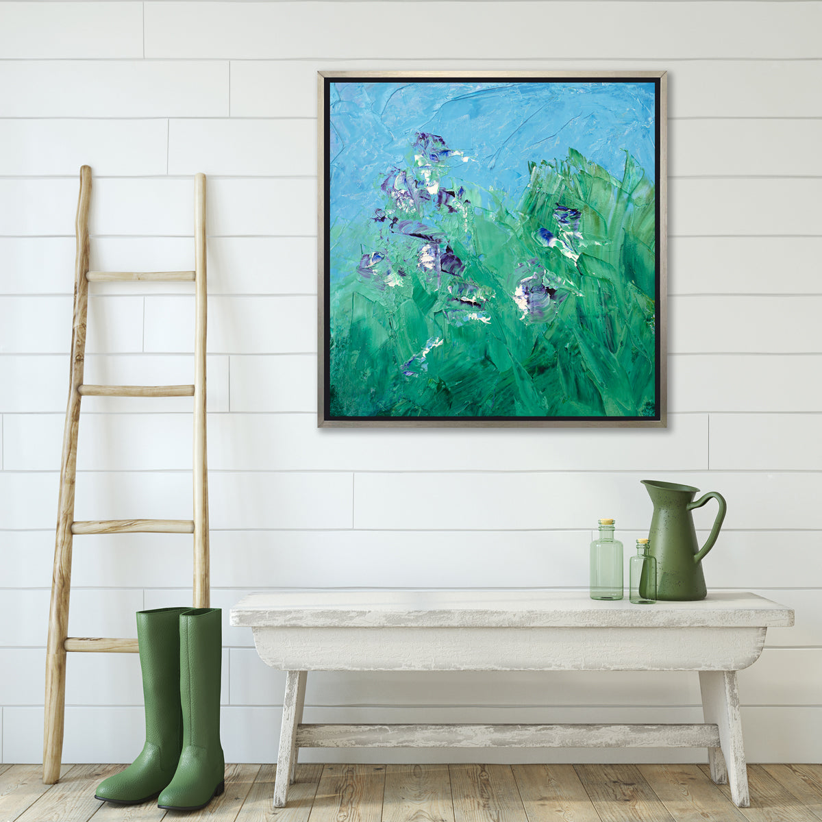 A blue, green, purple and white abstract landscape print in a silver floater frame hangs on a white wall above a whitewashed benched with two green bottles, a green pitcher, a pair of green rubber boots and a wood ladder.