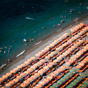 A side view aerial photograph of a beach club in the Amalfi Coast overlooking orange and yellow umbrellas and people relaxing in the ocean. 