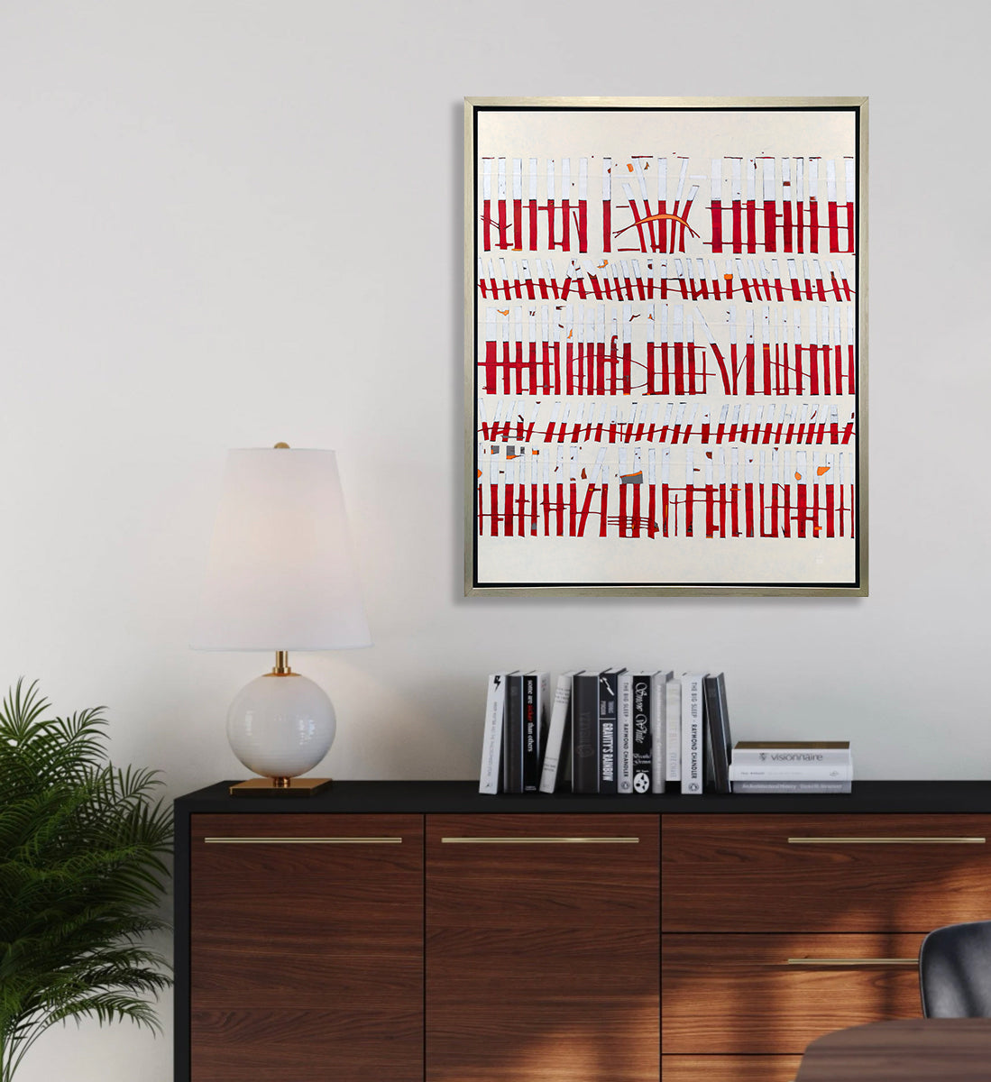 A beige, white and red abstract print in a silver floater frame hangs on a white wall above a dark wood and black cabinet with a stack of books, a white table lamp and a potted plant in the corner.