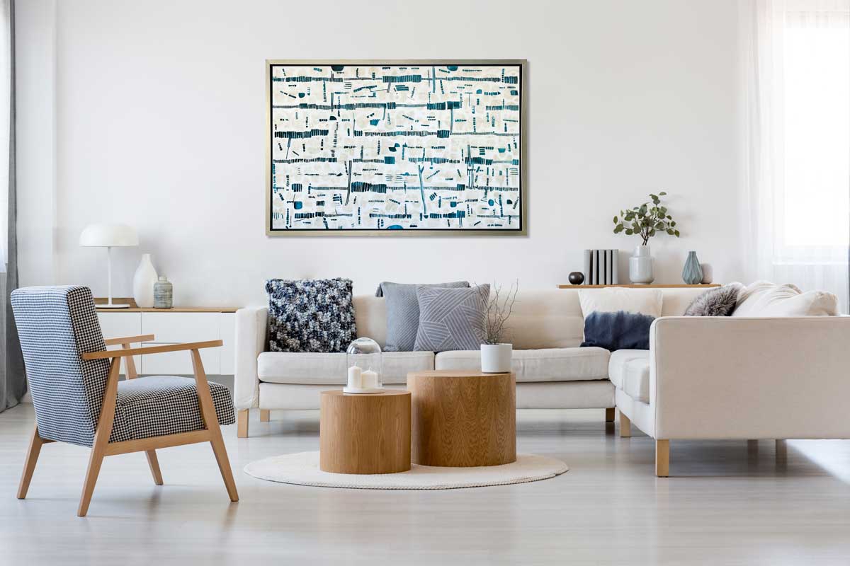A dark teal, white and beige abstract print in a silver floater frame hangs on a white wall above a white sectional sofa with blue and white printed pillows, two wooden solid round coffee tables and a blue and white checked chair.
